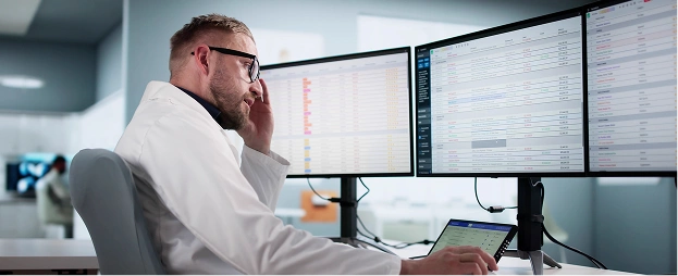 A person in a lab coat sits at a desk and works across two large monitors while holding a tablet