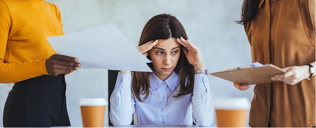 A woman sits at a desk holding her head while two people hand her papers. She looks stressed as work piles up around her