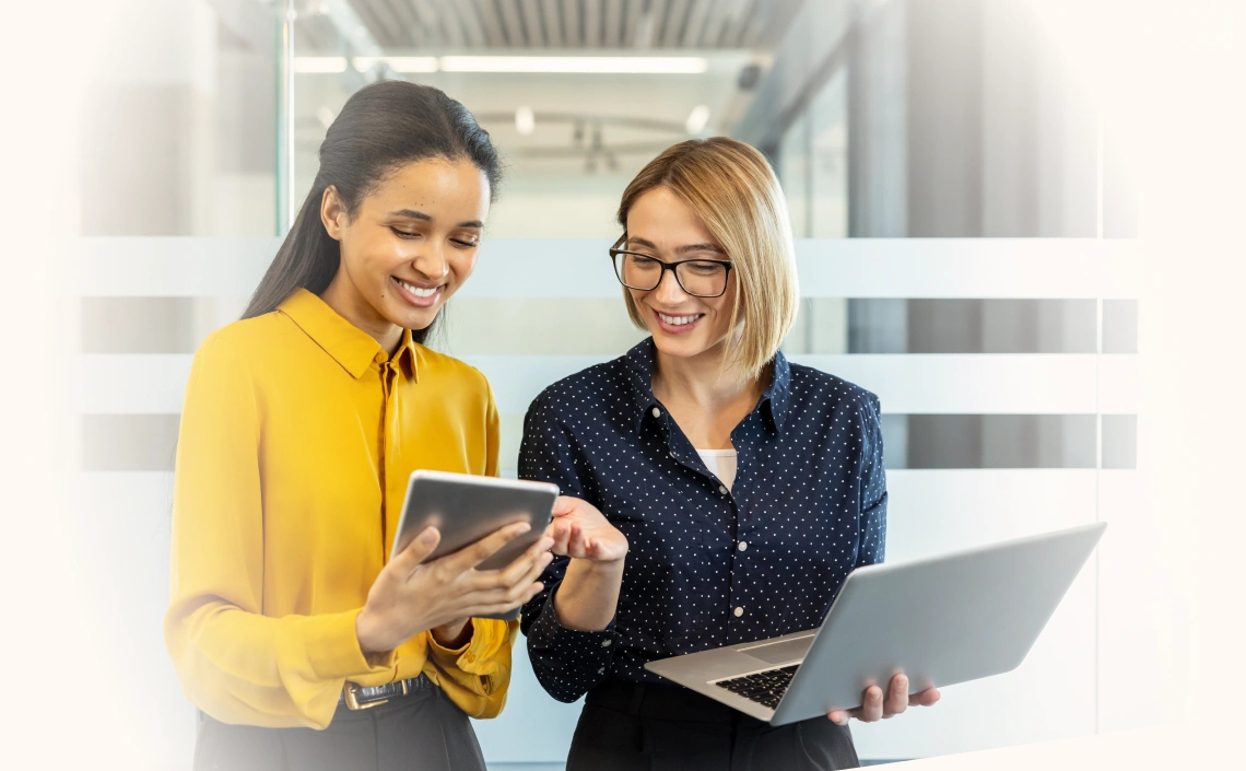 Two smiling businesswomen standing in an office, actively collaborating and discussing work using a laptop and a digital tablet, reflecting teamwork and modern communication